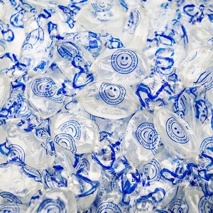 May include: A close-up of a pile of clear, individually wrapped candies. The wrappers are blue and white with the text "No Artificial Colours" and a smiley face.