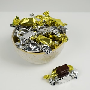 May include: A ceramic bowl filled with wrapped candies in gold and silver foil. A few candies are outside the bowl. The candies are dark brown and rectangular. The bowl has a floral pattern.