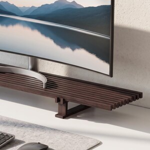May include: A dark brown wooden monitor stand featuring a slatted surface design supported by two rectangular legs. The stand holds a wide curved computer monitor displaying a mountain landscape reflection, with a grey keyboard and mouse on a desk mat.