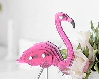May include: A pink flamingo sculpture made of layered acrylic, standing on a clear acrylic base. The sculpture is on a white surface with a white vase of white flowers in the background.