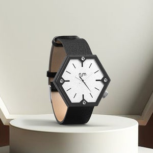 May include: A black hexagonal watch with a white face and black hands. The watch has a black leather band and is set on a white pedestal.