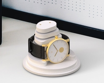 May include: A gold watch with a white face and a brown leather band is displayed on a white stand. The watch has a small gold circle in the center of the face.