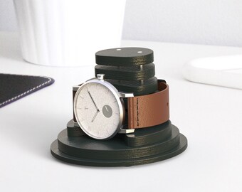 May include: A dark green watch stand with a silver watch and a brown leather band. The watch stand is made of stacked circles.