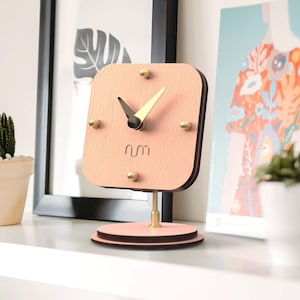 May include: A square, light pink wooden clock with gold-colored accents and hands. The clock sits on a gold stand and a round base. The clock is on a white shelf with a framed picture, magazines, and a small potted plant.