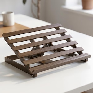 May include: A dark brown wooden laptop stand featuring a slatted design with horizontal bars. The stand has an adjustable angled frame connected by small metal hinges and a front ledge to support a device, resting on a flat surface.