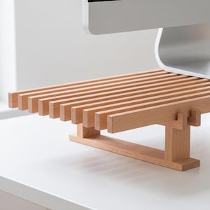 Op de afbeelding: Een monitorstandaard van lichtgekleurd hout met een lattenoppervlak bestaande uit parallelle horizontale staven. De standaard wordt ondersteund door twee verticale poten die verbonden zijn door een horizontale basisbalk voor een verhoogd platform.