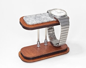 Op de afbeelding: Een houten horloge standaard met een grijs vilt kussen om het horloge te beschermen. De standaard heeft drie metalen staven om het horloge op zijn plaats te houden.