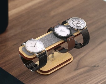 Op de afbeelding: Een houten horlogehouder met drie vakken voor horloges. De houder is gemaakt van licht en donker hout en heeft een grijze viltvoering. Drie horloges met verschillende gekleurde banden worden op de houder gepresenteerd.