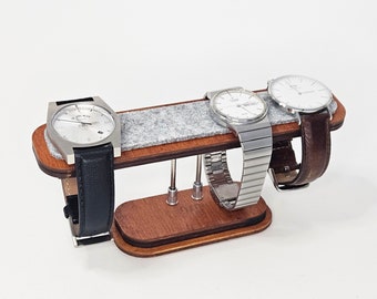 Op de afbeelding: Een houten horlogehouder met drie vakken voor horloges. De houder is gevoerd met grijs vilt en is gemaakt van donker hout. Drie horloges worden op de houder gepresenteerd, twee met zilveren banden en een met een zwarte band.