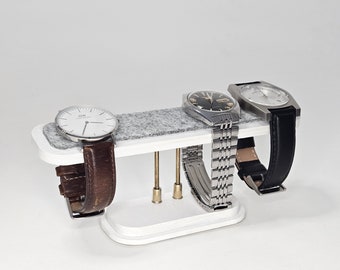 May include: White watch stand with three watches. Two watches have silver bands and one has a brown band. The stand has a gray felt lining.