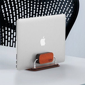 May include: A silver laptop stand with a wooden base, holding a silver laptop with an Apple logo. The laptop is on a white desk with a black keyboard and a black office chair in the background.