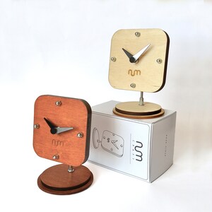May include: Two wooden clocks with a simple design. The clocks are square and have a black hour and minute hand. The clock face is white with black numbers and the word "nm" in the center. The clocks are on a white surface.