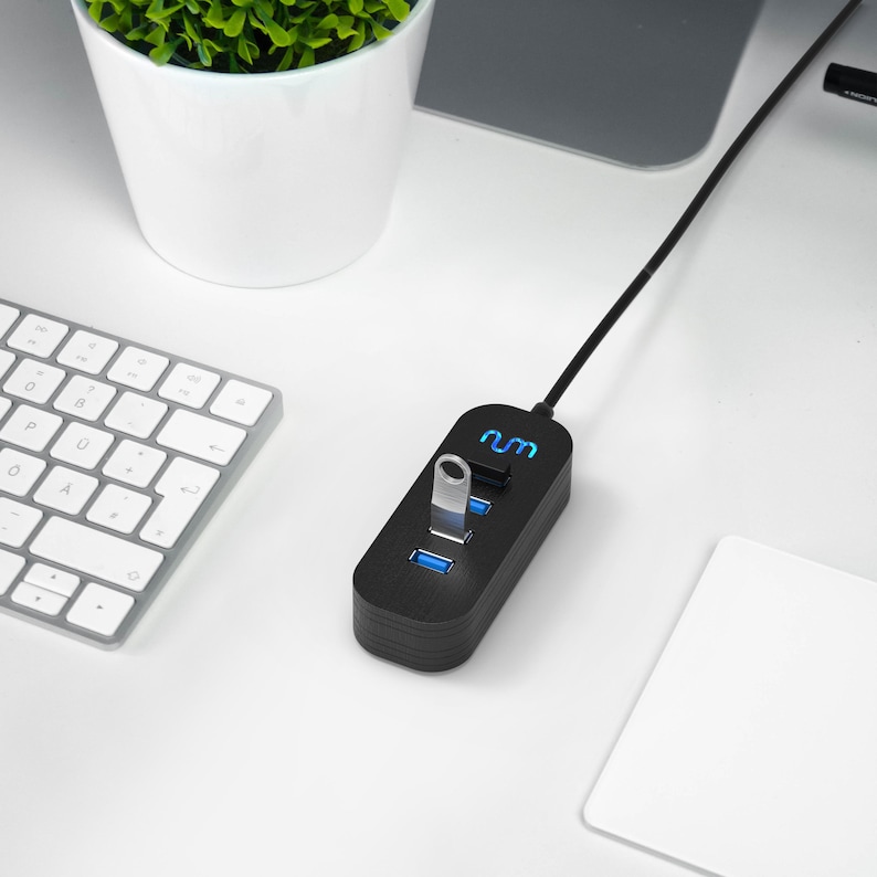 May include: A black USB hub with three ports and a black cable. The hub has a blue light and the text "num" on it. A silver USB drive is plugged into one of the ports.