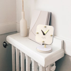 May include: White analog clock with gold hands and accents on a white stand. The clock face has the word "num" printed on it.