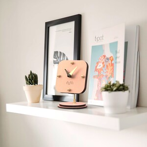 May include: A pink wooden clock with gold accents and black hands. The clock is on a small wooden stand and is sitting on a white shelf with a potted cactus and a magazine.