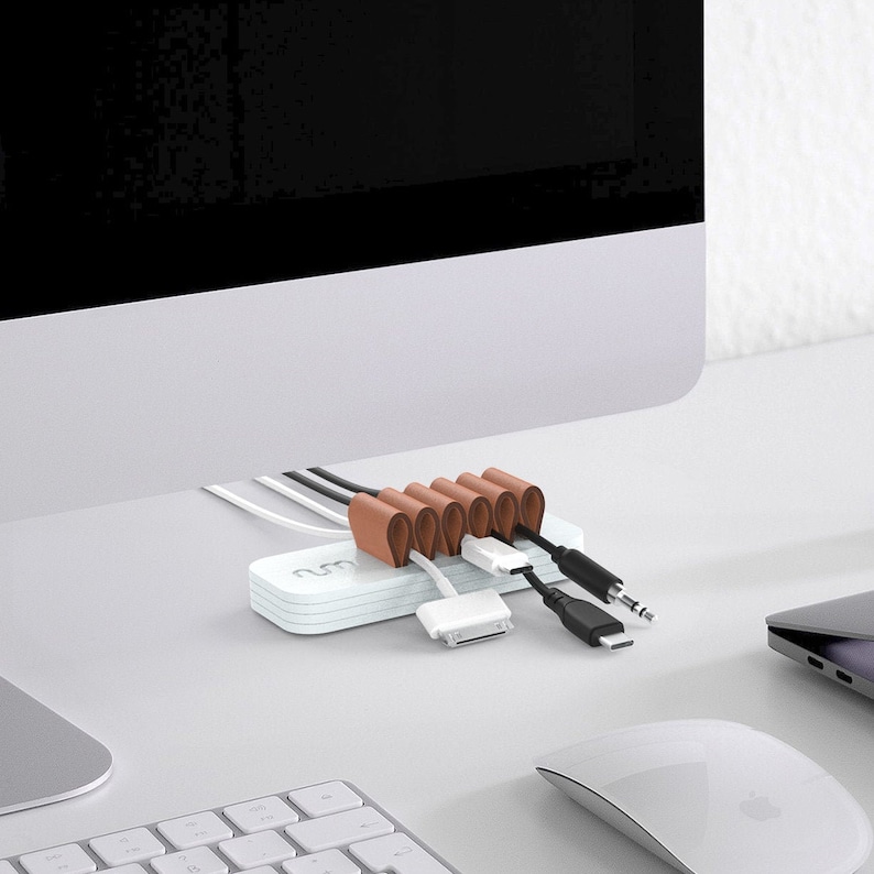 May include: A white cable organizer with brown leather straps to hold cords in place. The organizer is sitting on a white desk with a computer monitor, keyboard, and mouse.