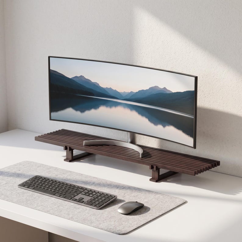 May include: A dark brown wooden monitor stand featuring a slatted surface design supported by two rectangular legs. The stand holds a wide curved computer monitor displaying a mountain landscape reflection, with a grey keyboard and mouse on a desk mat.