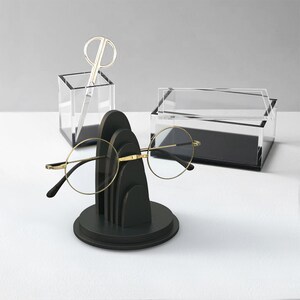 May include: A black wooden stand with a rounded top and three tiers, designed to hold eyeglasses. The stand is on a white surface, and a pair of gold-rimmed eyeglasses is resting on the stand.