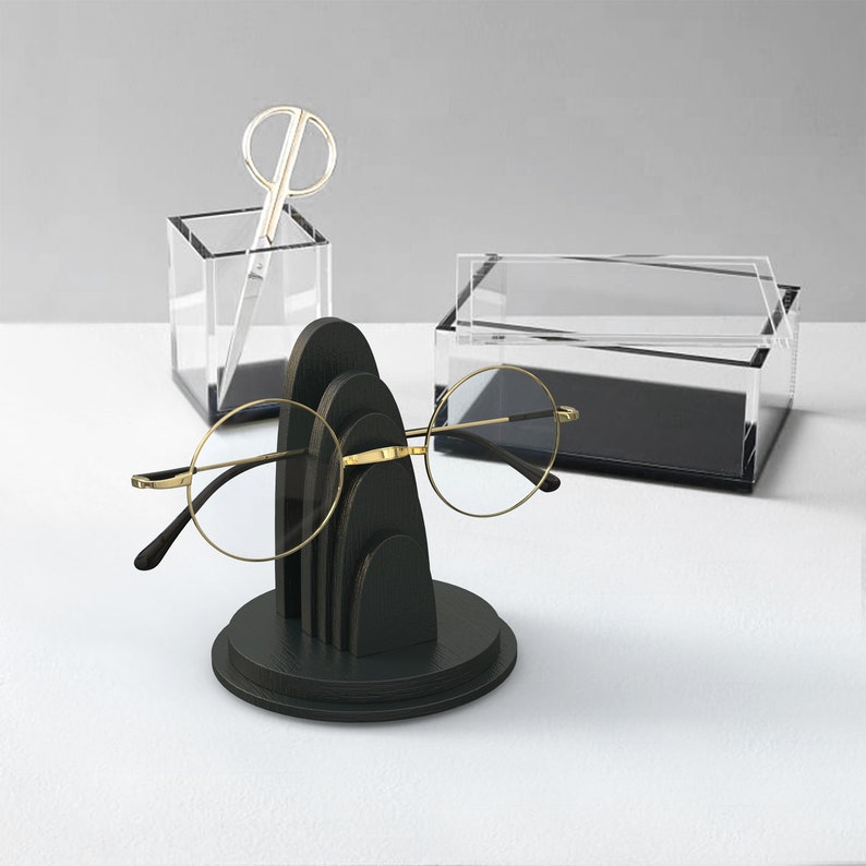 May include: A black wooden stand with a rounded top and three tiers, designed to hold eyeglasses. The stand is on a white surface, and a pair of gold-rimmed eyeglasses is resting on the stand.