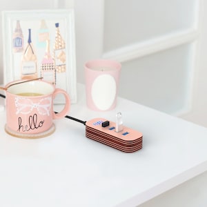 May include: A pink and brown USB hub with four ports and a blue light. The hub is connected to a laptop with a black cable. The hub is sitting on a white table.
