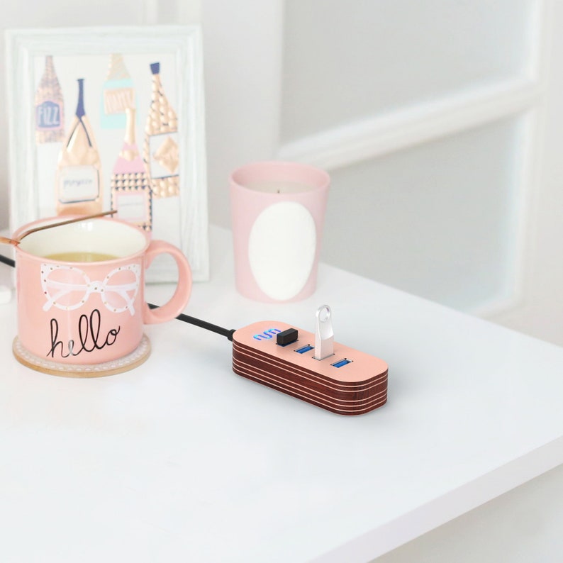 May include: A pink and brown USB hub with four ports and a blue light. The hub is connected to a laptop with a black cable. The hub is sitting on a white table.