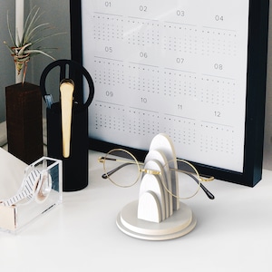 May include: White wooden glasses holder with a geometric design. The holder has three arches, each with a different size. The holder is on a white surface. A pair of gold-rimmed glasses is resting on the holder.