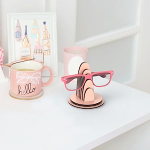 May include: Pink wooden glasses holder with a stack of five arches. A pair of pink glasses is resting on the holder. The holder is on a white surface.