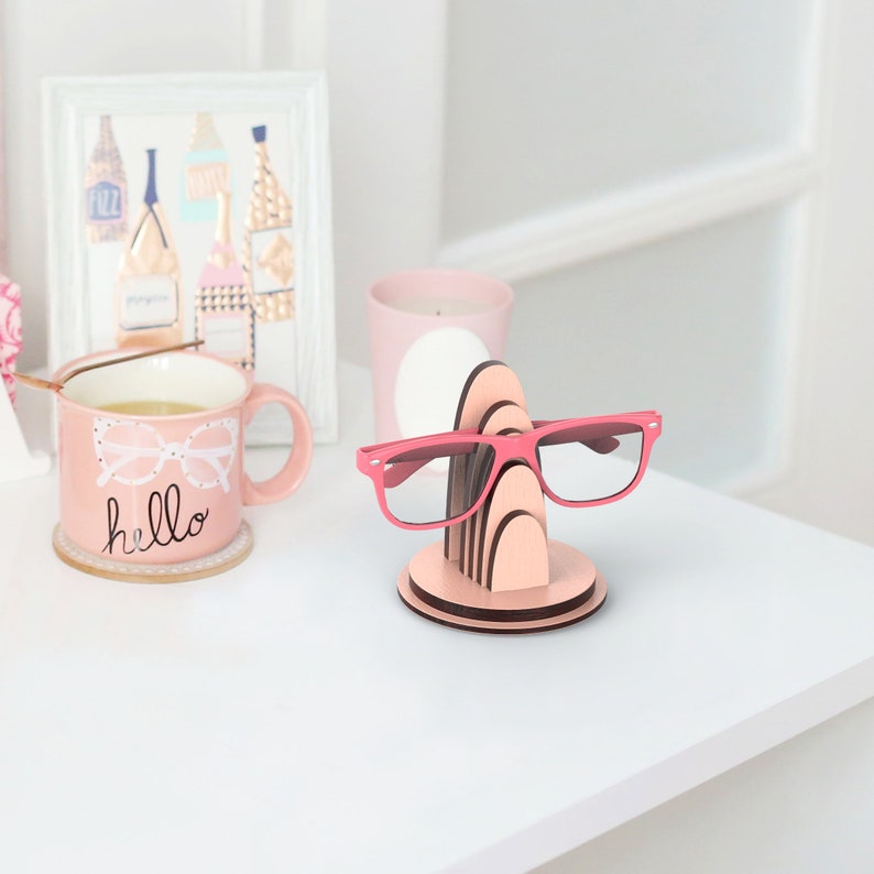 May include: Pink wooden glasses holder with a stack of five arches. A pair of pink glasses is resting on the holder. The holder is on a white surface.
