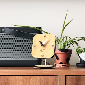May include: A wooden clock with a white face and black hands. The clock is on a stand and has the word "num" printed on the face. The clock is in front of a black speaker and a potted plant.