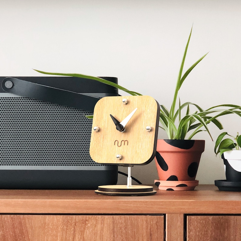 May include: A wooden clock with a white face and black hands. The clock is on a stand and has the word "num" printed on the face. The clock is in front of a black speaker and a potted plant.
