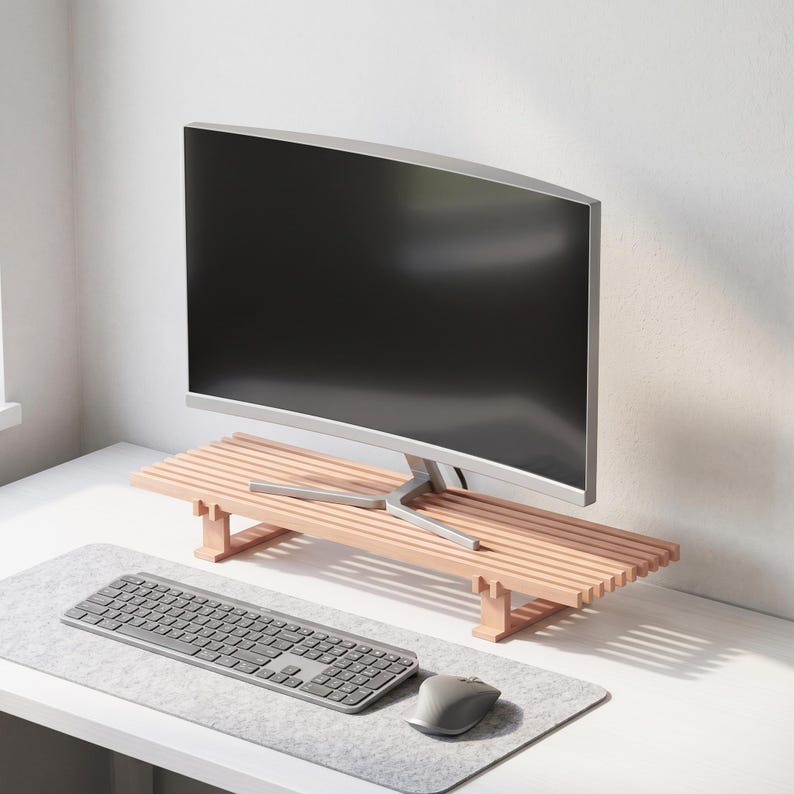 Op de afbeelding: Een monitorstandaard van lichtgekleurd hout met een latjesoppervlak en twee stevige steunpoten. De standaard staat op een bureau en draagt een gebogen computermonitor, met een grijs toetsenbord en een computermuis op een vilten mat eronder.