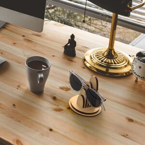 May include: A wooden sunglasses stand with a modern design. The stand is made of light brown wood with a darker brown accent. The stand is holding a pair of sunglasses with black lenses.