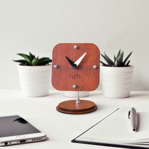 May include: A wooden desk clock with a square face and black hands. The clock is on a wooden stand and has a white background with the letters "rm" in the center.