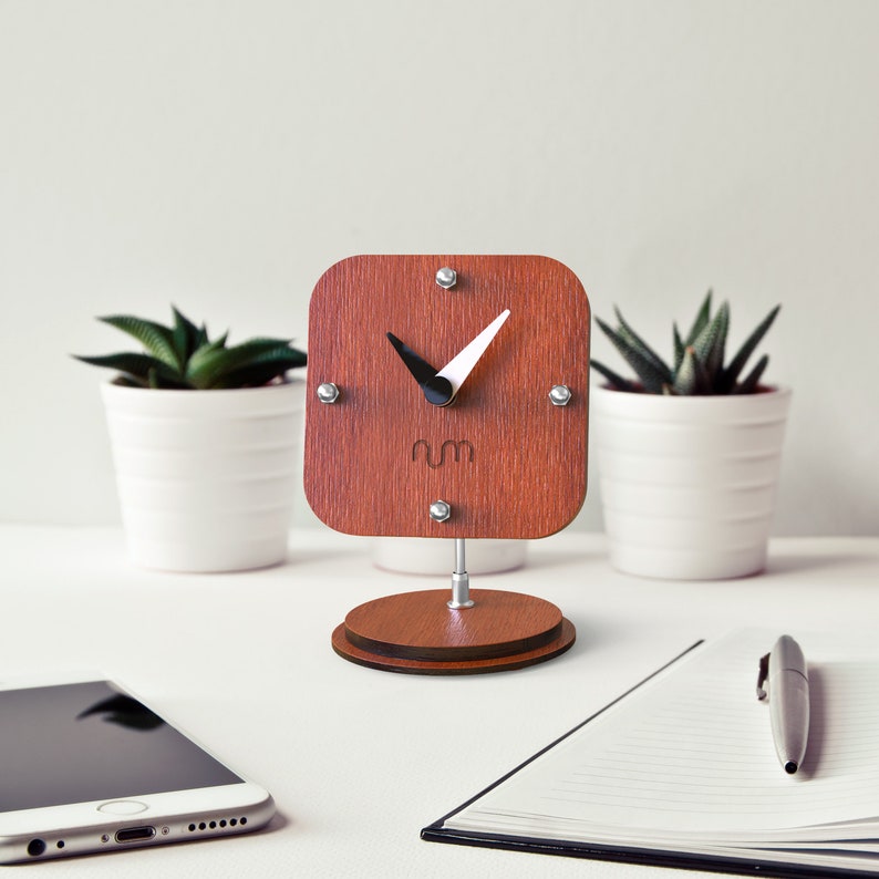 May include: A wooden desk clock with a square face and black hands. The clock is on a wooden stand and has a white background with the letters "rm" in the center.