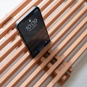 Op de afbeelding: Een lichtgekleurde houten standaard met een lattenontwerp met parallelle staven. Een zwarte smartphone staat rechtop in de standaard en toont de tijd 10:09 en de datum Sali, 10 Aralik op het scherm. De standaard heeft een rasterachtige structuur.