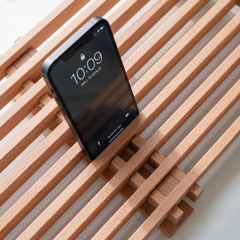 Op de afbeelding: Een lichtgekleurde houten standaard met een lattenontwerp met parallelle staven. Een zwarte smartphone staat rechtop in de standaard en toont de tijd 10:09 en de datum Sali, 10 Aralik op het scherm. De standaard heeft een rasterachtige structuur.