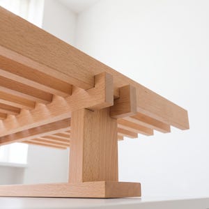 Op de afbeelding: Een lichtgekleurde houten bank met een zitting van latten en zichtbare verbindingen. De structuur bestaat uit horizontale houten latten die worden ondersteund door een centrale verticale paal en een platte rechthoekige basis van natuurlijk hout.