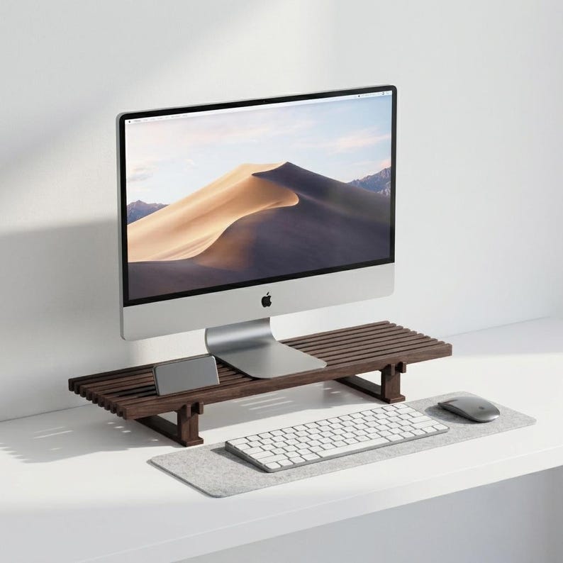 Op de afbeelding: Een donkerbruine houten monitorstandaard met een lattenoppervlak, een rechthoekige vorm en vier korte poten. De standaard ondersteunt een zilveren computermonitor, een kleine smartphone, een wit toetsenbord en een grijze computermuis.