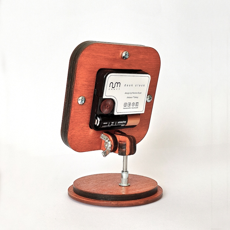 May include: A wooden desk clock with a brown wood frame and a white face. The clock is on a stand and has a silver metal knob. The clock face has the text "nm craft desk clock" and "design by Nurhan Ayaz Ankara / Turkey".