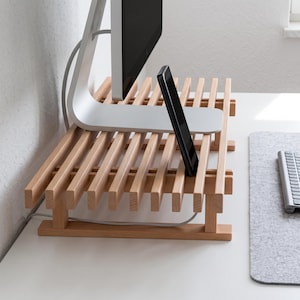 Op de afbeelding: Een monitorstandaard van lichtgekleurd hout met een lattenontwerp van horizontale balken. De standaard ondersteunt de zilveren voet van een computermonitor en houdt een zwarte smartphone rechtop tussen de latten. Het rust op vier kleine pootjes.