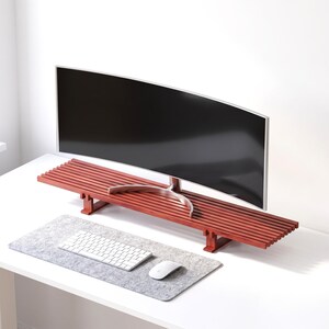 Op de afbeelding: Een roodbruine houten monitorstandaard met een lattenontwerp, een rechthoekig blad en vier korte poten. Een gebogen computermonitor staat op de standaard, terwijl een wit toetsenbord en een witte computermuis op een grijze vilten bureauonderlegger liggen.