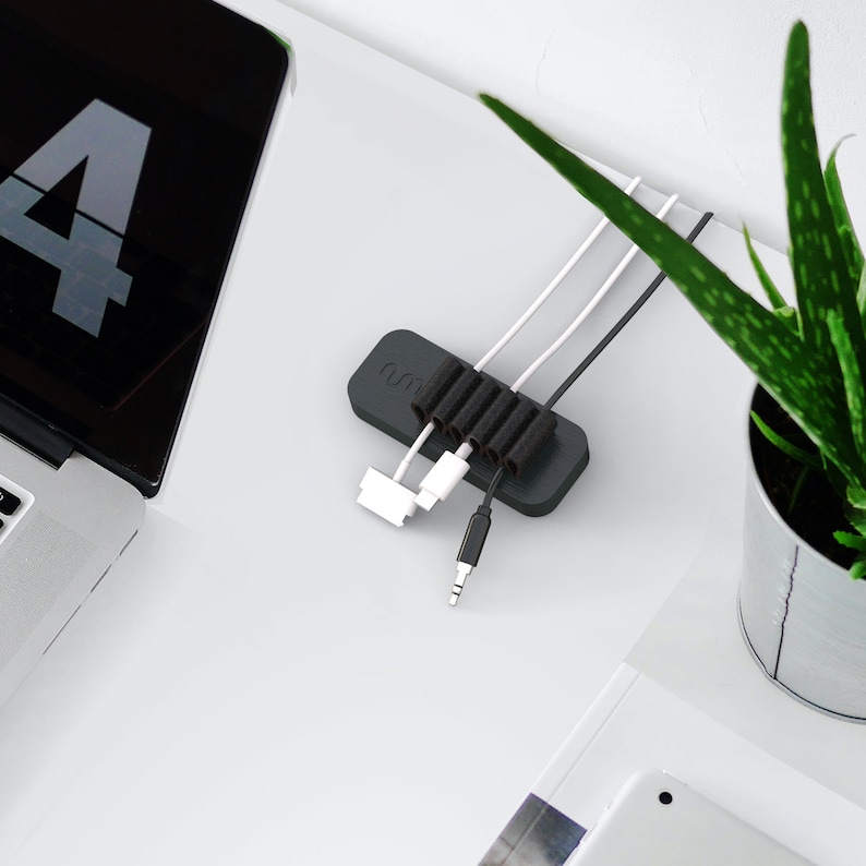 May include: A black cable organizer with six slots to hold cords and wires. The organizer has the word "num" printed on it. The organizer is on a white surface with a laptop and a potted plant.