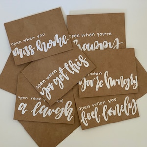 May include: A collection of brown paper envelopes with white handwritten text. The text includes phrases like "open when you miss home," "open when you get this," and "open when you're hungry." The envelopes are arranged in a slightly overlapping pattern.