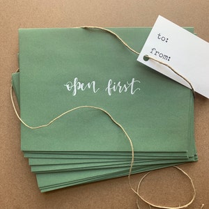 May include: A stack of green envelopes with the words "open first" in white script. A white tag with "to:" and "from:" is attached with twine. The twine is also wrapped around the envelopes.