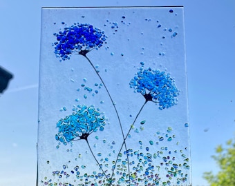 May include: A glass art piece featuring three blue and turquoise flowers with white stems on a clear background. The flowers are made of small glass pieces and are scattered with iridescent glitter.