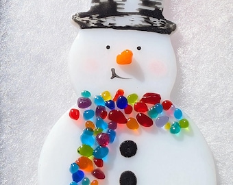 Könnte beinhalten: Schneemann-Ornament mit schwarzem Zylinder und buntem Perlschal. Der Schneemann hat eine orangefarbene Karottennase, rosige Wangen und drei schwarze Knöpfe. Ein rotes Band ist zum Aufhängen befestigt.