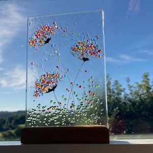 May include: A glass art piece featuring two colourful dandelion-like flowers with scattered glass beads in shades of green, blue, red, yellow, and orange. The piece is mounted on a wooden stand.