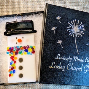Peut inclure: Ornement de bonhomme de neige en verre blanc avec un chapeau haut de forme noir et une écharpe de perles colorées. Le bonhomme de neige a un nez orange souriant et trois boutons noirs. Dans une boîte noire avec le texte "Lovingly Made By Loxley Chapel Glass".