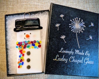 Könnte beinhalten: Ein weißer Schneemann-Glasornament mit schwarzem Zylinder und einem bunten Perlen-Schal. Der Schneemann hat eine lächelnde orangefarbene Nase und drei schwarze Knöpfe. In einer schwarzen Box mit dem Text "Lovingly Made By Loxley Chapel Glass".