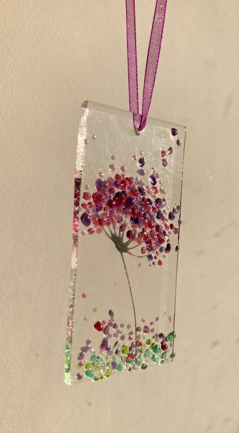 K&ouml;nnte beinhalten: Ein Glasornament mit einem lila und pinkfarbenen Blumendesign. Die Blume besteht aus kleinen Glasperlen und ist von kleineren gr&uuml;nen und lila Perlen umgeben.